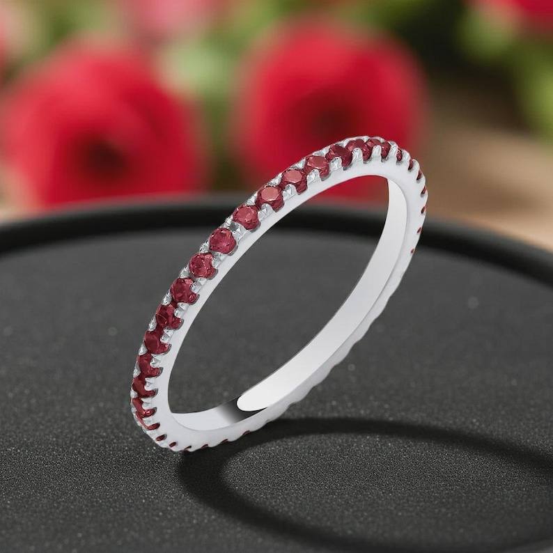 Pode incluir: Um anel de banda fina em tom prateado com uma fila cont&iacute;nua de pequenas pedras preciosas redondas de cor vermelho-escura engastadas ao longo da superf&iacute;cie exterior. O anel tem um acabamento met&aacute;lico polido e uma forma circular uniforme.