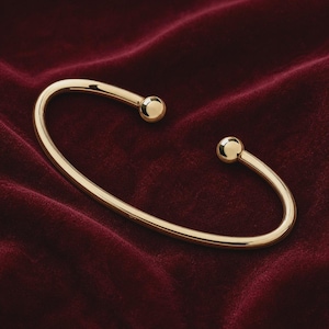 May include: A gold-coloured open cuff bracelet with rounded ball ends. The bracelet has a smooth, polished finish and is set against a deep red velvet background. The design is simple and elegant.
