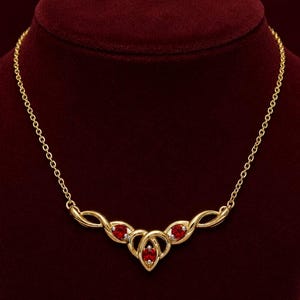May include: A gold necklace featuring a central pendant with three round, deep red gemstones set in polished gold metal. The pendant has an intricate, swirling design that connects to a delicate gold chain, creating a cohesive piece of jewellery.