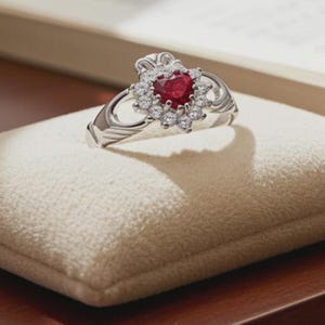 May include: A silver Claddagh ring with a heart-shaped ruby center stone, surrounded by small white gemstones. The ring rests on a small, cream-colored pillow. The ring's design features a heart, hands, and a crown.