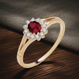 May include: A gold ring featuring a central, round, deep red gemstone surrounded by a halo of smaller, clear gemstones. The ring's band is a polished gold color, with a split design near the gemstone setting.