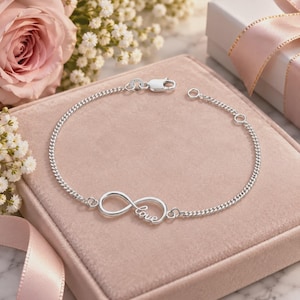 Könnte beinhalten: Ein silbernes Kettenarmband mit einem Unendlichkeitssymbol-Anhänger, in den das Wort love in Schreibschrift eingearbeitet ist. Das Armband verfügt über einen Karabinerverschluss und eine kurze Verlängerungskette mit zwei runden Gliedern.