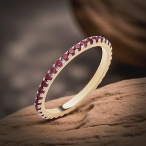 Könnte beinhalten: Ein dünner Goldring mit einer durchgehenden Reihe kleiner, runder, dunkelroter Edelsteine, die in silberfarbenen Krappen am äußeren Rand gefasst sind. Der Ring hat eine polierte Goldinnenseite und ein glattes, schmales Profil.