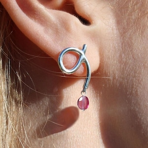 May include: Silver dangle earrings with a pink gemstone. The earrings feature a curved, abstract design.