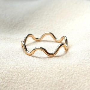 May include: A gold-colored ring with a wavy design. The ring has a continuous, flowing pattern, resembling a series of gentle waves. The ring is set against a soft, neutral background, highlighting its simple elegance. The ring is a piece of jewelry.