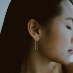 May include: A close-up of a person's ear with a silver rectangle earring. The earring is simple and minimalist.