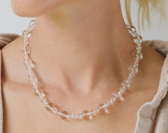 May include: A clear quartz bead necklace with a delicate design. The necklace is composed of round, transparent beads of varying sizes. The image also shows a pair of gold earrings and a beige button-down shirt.