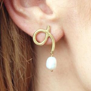 May include: Gold-toned, abstract-shaped earrings with a dangling white pearl. The earrings have a unique, swirling design and a small, round pearl at the bottom. The earrings are shown on a person's ear.