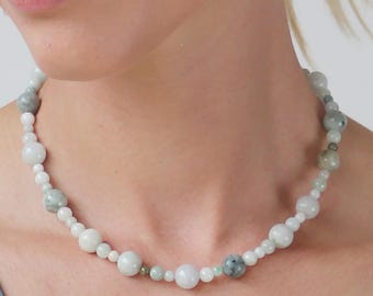 May include: A beaded necklace with white and green stones, worn with silver drop earrings. The woman is wearing a teal top. The necklace is a piece of jewellery.