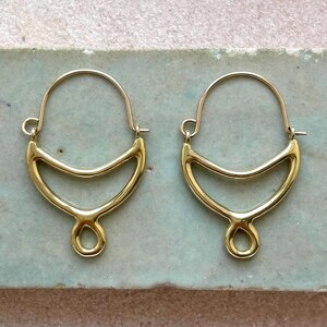 May include: A pair of gold-colored hoop earrings. Each earring features a curved, open design with a rounded top and a teardrop-shaped bottom. The earrings are displayed on a light blue and tan surface.