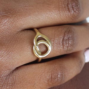 May include: A gold-colored ring featuring a knot design. The ring is made of a shiny, metallic material and has a smooth surface. The knot is the focal point, creating a unique and stylish accessory. The ring is shown on a person's finger.