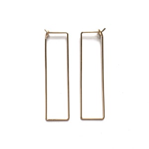 May include: A pair of gold-coloured rectangular hoop earrings. The earrings have a simple, minimalist design, with a long, thin rectangular shape. The earrings are displayed on a white background.