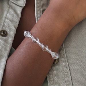 May include: A clear quartz crystal bracelet with varying sized, round beads. The bracelet is strung together and worn on the wrist. The background includes a light green shirt.