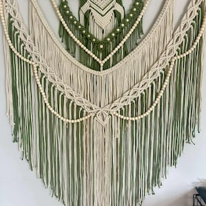 May include: A macrame wall hanging in green and cream. The design features intricate knotting patterns, wooden beads, and long fringe. The piece is suspended from a natural wooden dowel, creating a bohemian-inspired decorative element for a home.