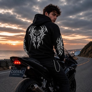 May include: A black hooded sweatshirt featuring a white tribal-style graphic print on the back and sleeves. The hoodie is worn with black trousers that have a silver chain attached to the pocket. The garment is displayed on a motorcycle with a number plate reading CARNAC BIKERS.