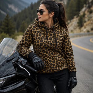 May include: A long-sleeved hooded sweatshirt featuring a brown and black floral pattern, black ribbed cuffs, and a black drawstring. The garment is paired with black trousers and black protective motorcycle gloves while leaning on a black motorcycle.