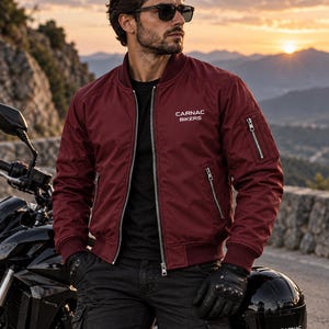 Può includere: Un giubbotto bomber bordeaux con chiusura frontale a zip, colletto e polsini a coste e una tasca multiuso con zip sulla manica sinistra. Il testo CARNAC BIKERS è stampato in bianco sulla parte superiore sinistra del petto.