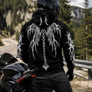 May include: A black hooded sweatshirt featuring a white, intricate, sharp-edged graphic design extending from the hood down the back and along the sleeves. The hoodie is worn with dark denim jeans and black gloves, paired with a black motorcycle helmet.