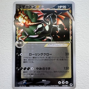 May include: A Charizard Pokémon trading card with a black and orange dragon illustration. The card has Japanese text and the HP is 90. The card is silver with a gold border and has the number 052/068.