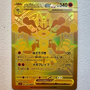 May include: A gold Pokémon trading card with a holographic effect, featuring a detailed illustration of a character. The card has Japanese text and displays the character's name and attack details. The card's design is intricate and eye-catching.