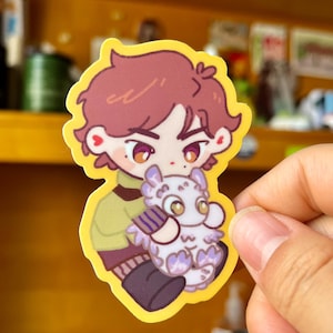 May include: A sticker featuring a cartoon character with brown hair, wearing a green and brown shirt, holding a small, purple and white creature with yellow eyes.