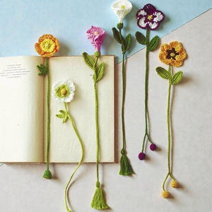 May include: A collection of handmade crochet flower bookmarks in various colors, including yellow, white, pink, and purple. Each bookmark features a detailed flower head, green leaves, and a long green stem with a tassel. The bookmarks are placed in an open book.