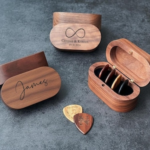 Op de afbeelding: Houten gitaar plectrumdozen met gepersonaliseerde gravure. Eén doos is open en toont plectrums. Twee extra dozen zijn gesloten, met namen en een oneindigheidssymbool gegraveerd op de deksels. Twee plectrums zijn zichtbaar.