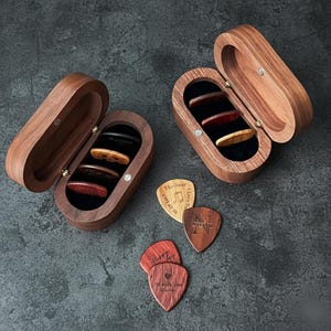 Könnte beinhalten: Zwei offene Gitarrenplektrum-Etuis aus Holz mit Plektren darin. Die Etuis sind oval und aus dunklem Holz gefertigt. Mehrere Gitarrenplektren sind um die Etuis herum verstreut, einige mit Text und Designs.