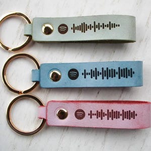 Könnte beinhalten: Drei Leder-Schlüsselanhänger in Pastellfarben: Hellgrün, Blau und Rosa. Jeder Schlüsselanhänger hat einen goldfarbenen Ring und ein Spotify-Code-Design. Die Schlüsselanhänger sind mit einem goldenen Knopf befestigt.
