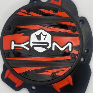 Peut inclure: Composant imprimé en 3D noir et rouge avec les lettres blanches « KYM ». Le design présente un emblème central et des motifs en forme d'éclair. Il comporte plusieurs trous et des éléments rouges en saillie.