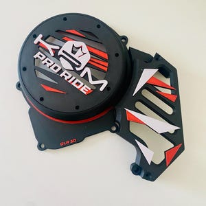 Peut inclure: Cache moteur de moto noir et rouge avec un logo "KTM Pro Ride" blanc et un graphique de couronne. Le cache a un motif géométrique noir et blanc sur le côté et indique "GLR 30" en rouge.