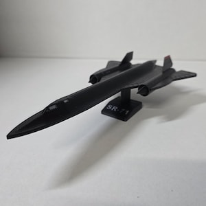Puede incluir: Maqueta negra de un avión SR-71, con un diseño elegante y alas. El modelo se apoya en un soporte negro con la inscripción "SR-71". Detalles en rojo en la cola. Una miniatura detallada.