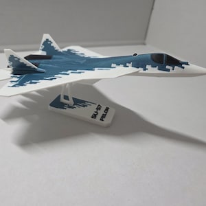 May include: A scale model of an SU-57 Felon fighter jet featuring a white and blue digital camouflage pattern. The aircraft is mounted on a white stand with the text SU-57 FELON printed on the base, showcasing a sleek aerodynamic design.