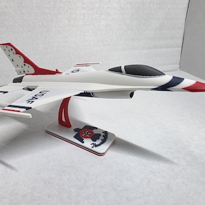 Puede incluir: Un modelo de avión blanco con detalles rojos y azules, incluido el texto "USAF" en las alas. El modelo se exhibe en un soporte con las palabras "Thunderbirds" y un logotipo. La cola presenta un patrón rojo y blanco.
