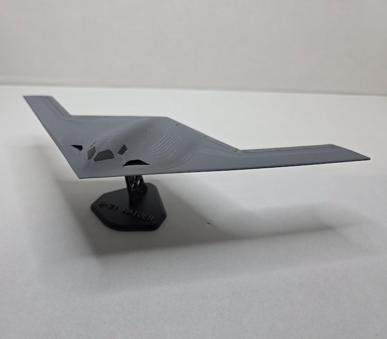 May include: A grey B-2 Spirit stealth bomber model, displayed on a black base. The model showcases a unique flying wing design with detailed surface features. The base has the text "B-21 RAIDER".