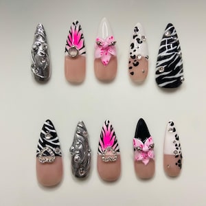 Pode incluir: Um conjunto de dez unhas artificiais com vários designs. As unhas apresentam uma mistura de padrões, incluindo listras de zebra, manchas de leopardo e detalhes florais. Algumas unhas são adornadas com strass e enfeites metálicos. A paleta de cores inclui rosa, preto, branco e nude.