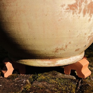 May include: A large terracotta pot with a worn, mottled surface sits on three terracotta supports. The pot is resting on a bed of moss and dirt.
