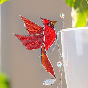 Peut inclure: Décoration d'oiseau cardinal en vitrail rouge, avec une tige en fil d'argent et des cristaux transparents. L'oiseau est posé sur le bord d'un pot en céramique blanc, près de feuilles vertes.