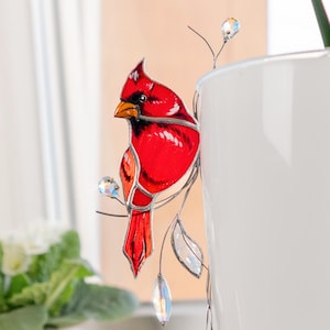 May include: A stained glass cardinal bird decoration in vibrant red, perched on a branch with clear crystal accents. The bird is set against a window, with a white flower pot and a white ceramic pot in the background.