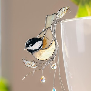 May include: A stained glass chickadee bird decoration with a black head, white cheeks, and tan body, perched on a delicate wire branch with crystal accents. The bird is near a white plant pot with green leaves.