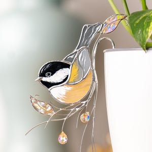 Op de afbeelding: Een vogel van gekleurd glas met zwarte, witte en gele accenten, die op een tak zit met kristallen accenten. De vogel hangt aan een witte keramische pot.