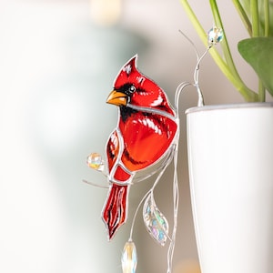 May include: A stained glass cardinal perched on a silver wire branch with iridescent crystal accents. The cardinal is red with a black beak and black eyes. The branch is attached to a white ceramic pot.