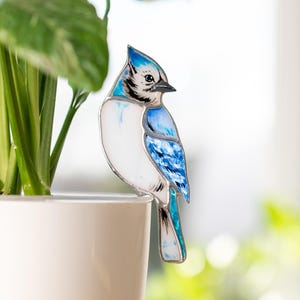 以下が含まれることがあります： 白と青のアクセントが施された、装飾用の青い鳥の飾り。鳥は白い植木鉢の端に止まっており、緑の葉の隣にあります。鳥は詳細なデザインで、ガラスまたは同様の素材で作られています。