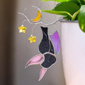May include: A stained glass bat ornament with a black body, purple and pink wings, and a pink face. The bat hangs from a silver branch with a yellow crescent moon and three yellow star charms. The ornament is placed in front of a green plant in a white pot.