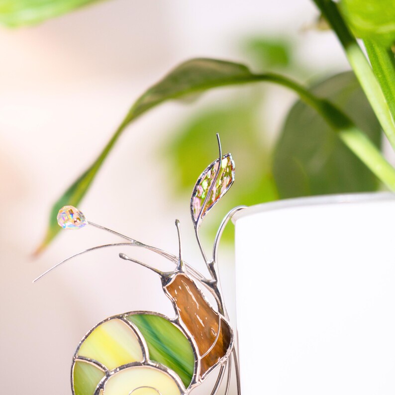 Detailed view of stained glass snail ornament with green shell and amber body attached to plant pot. Hanging crystal beads create soft rainbow reflections.
