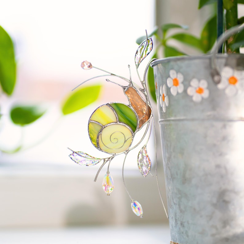 Stained glass snail plant decoration attached to metal bucket planter with daisy design. Green spiral shell and iridescent glass accents with crystal beads create charming garden style decor.