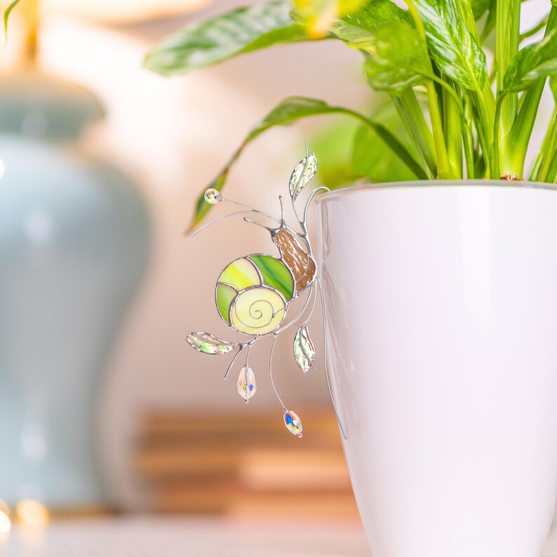 Handmade stained glass snail ornament clipped to white flower pot in cozy interior. Translucent green shell and crystal drops reflect warm light, elegant plant decor gift.