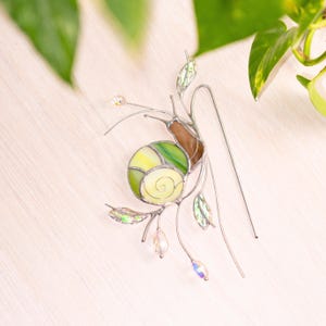 Handmade stained glass snail suncatcher with green and yellow shell placed on light wooden surface, detailed glass art with metal wire leaves and hanging crystals, unique plant decor accessory.