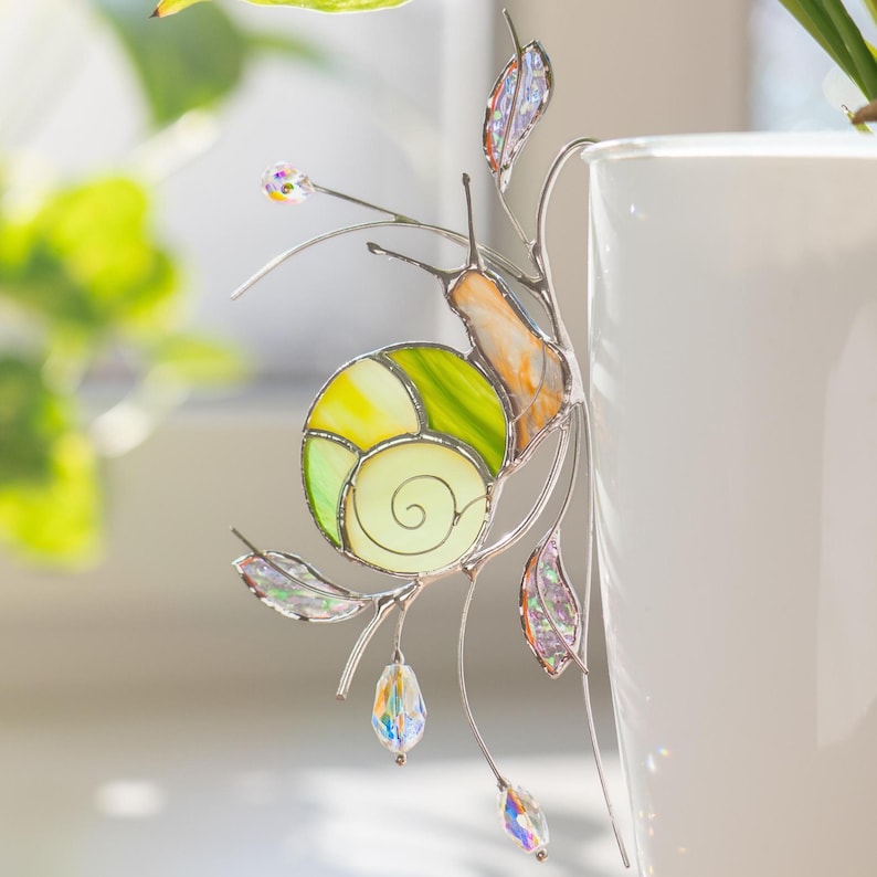 Handmade stained glass snail plant pot decor clipped to white planter near green houseplant. Green and yellow glass shell with crystal drops sparkles in sunlight, unique whimsical indoor plant decoration.