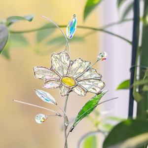 May include: A stained glass flower with a yellow center and iridescent petals. The flower has a silver metal stem and leaves. The design includes clear, colorful glass accents, creating a decorative floral piece.
